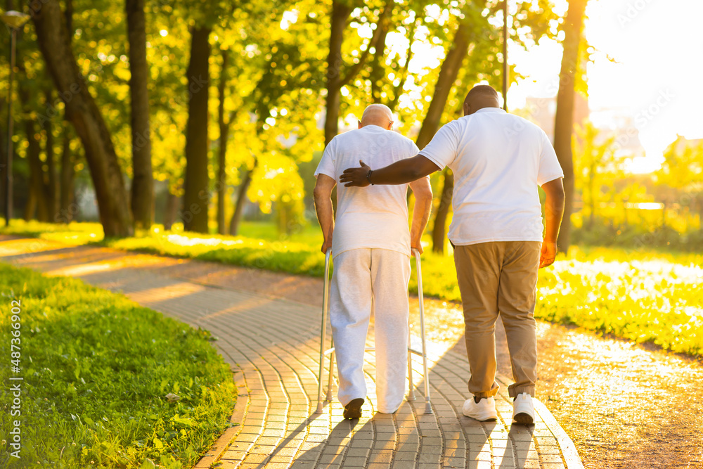 a care worker helping an elderly man with a walking frame walk along a path