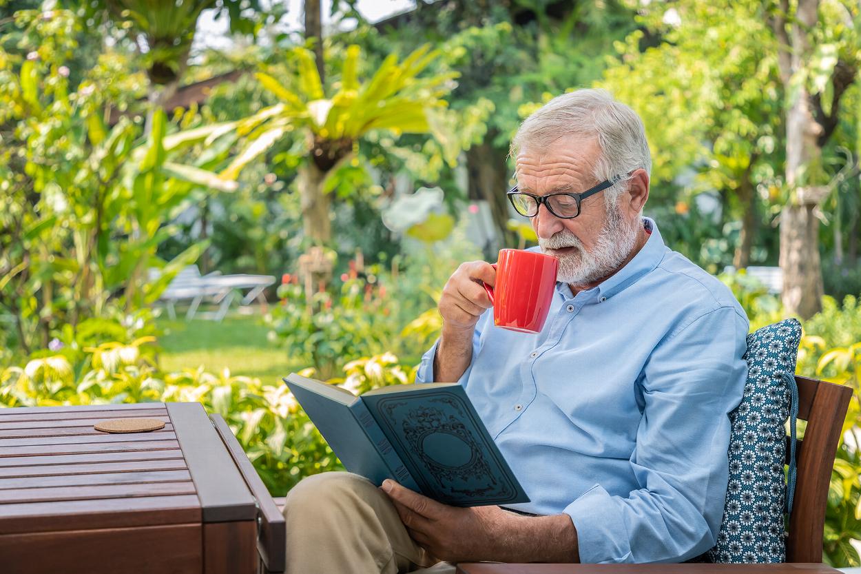 an old man reading a book in a garden while drinking from a red mug